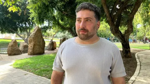 BBC Noam Glukhovsky stands outside in a park wearing a beige T-shirt