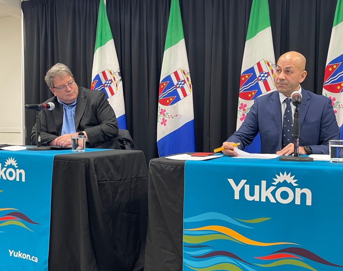 Two men sit behind tables with microphones and 'Yukon' banners.